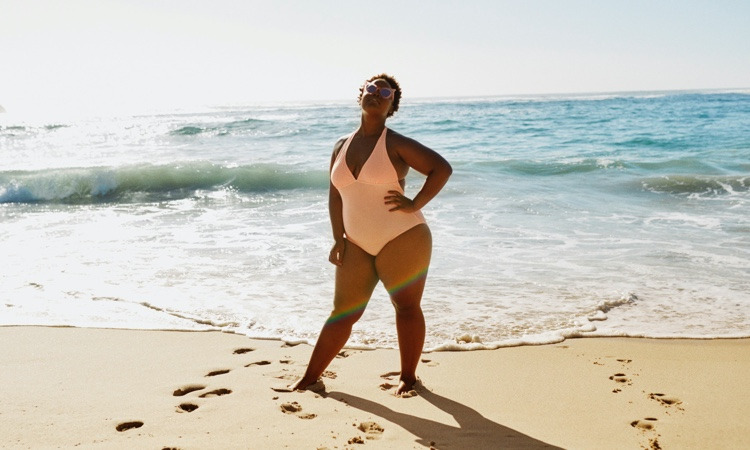 Plus sized woman in sunglasses and peach swimming costume stands on a beach.
